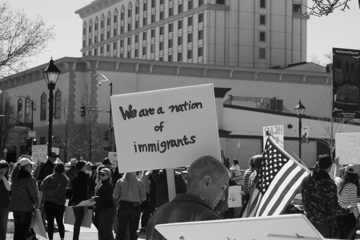 Black and white image of protester holding we are a nation of immigrants sign with American flag.