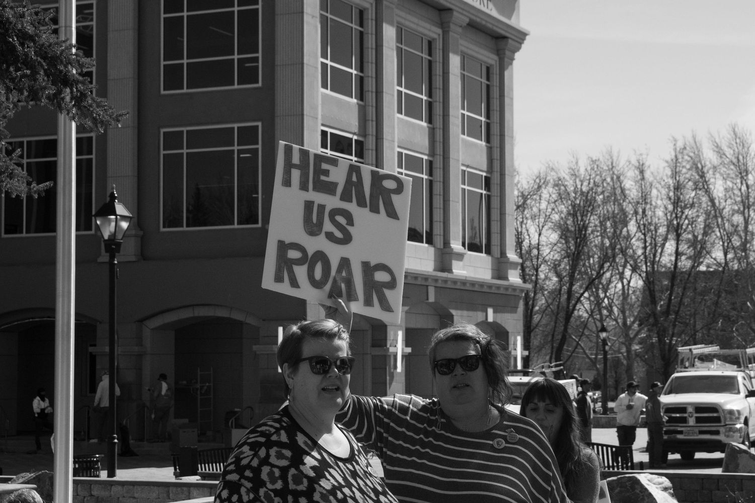 Two women pose holding a Hear Us Roar sign at an outdoor protest rally in an urban setting.
