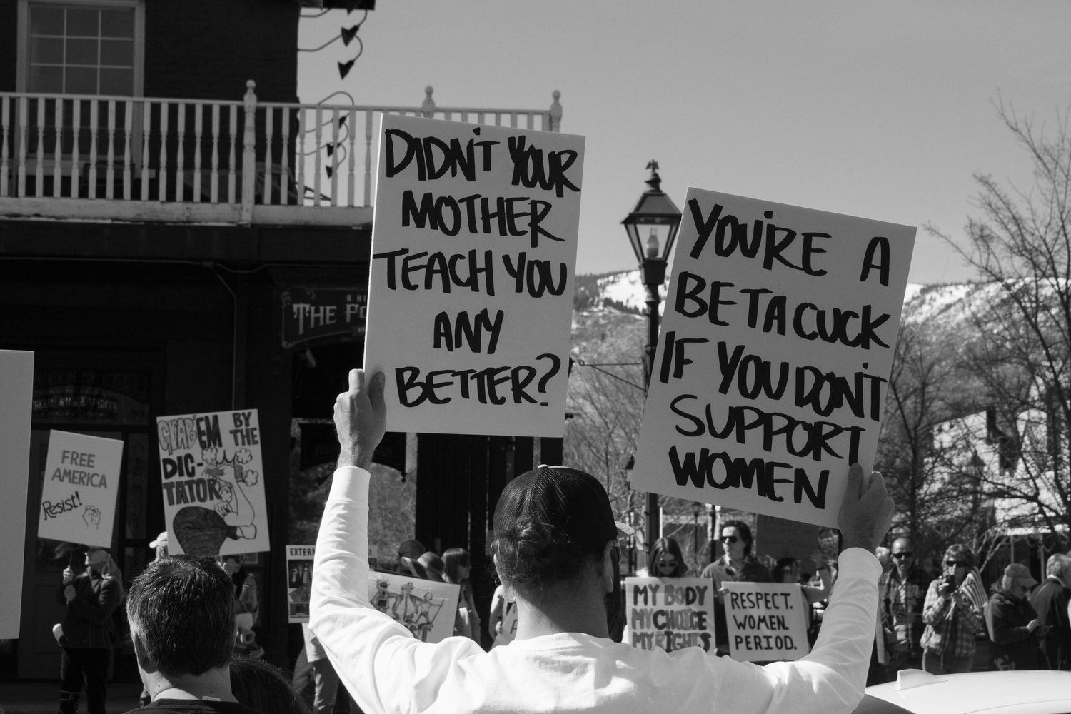 Two protesters hold signs reading Didnt Your Mother Teach You Any Better and Beta Cuck at a rally.