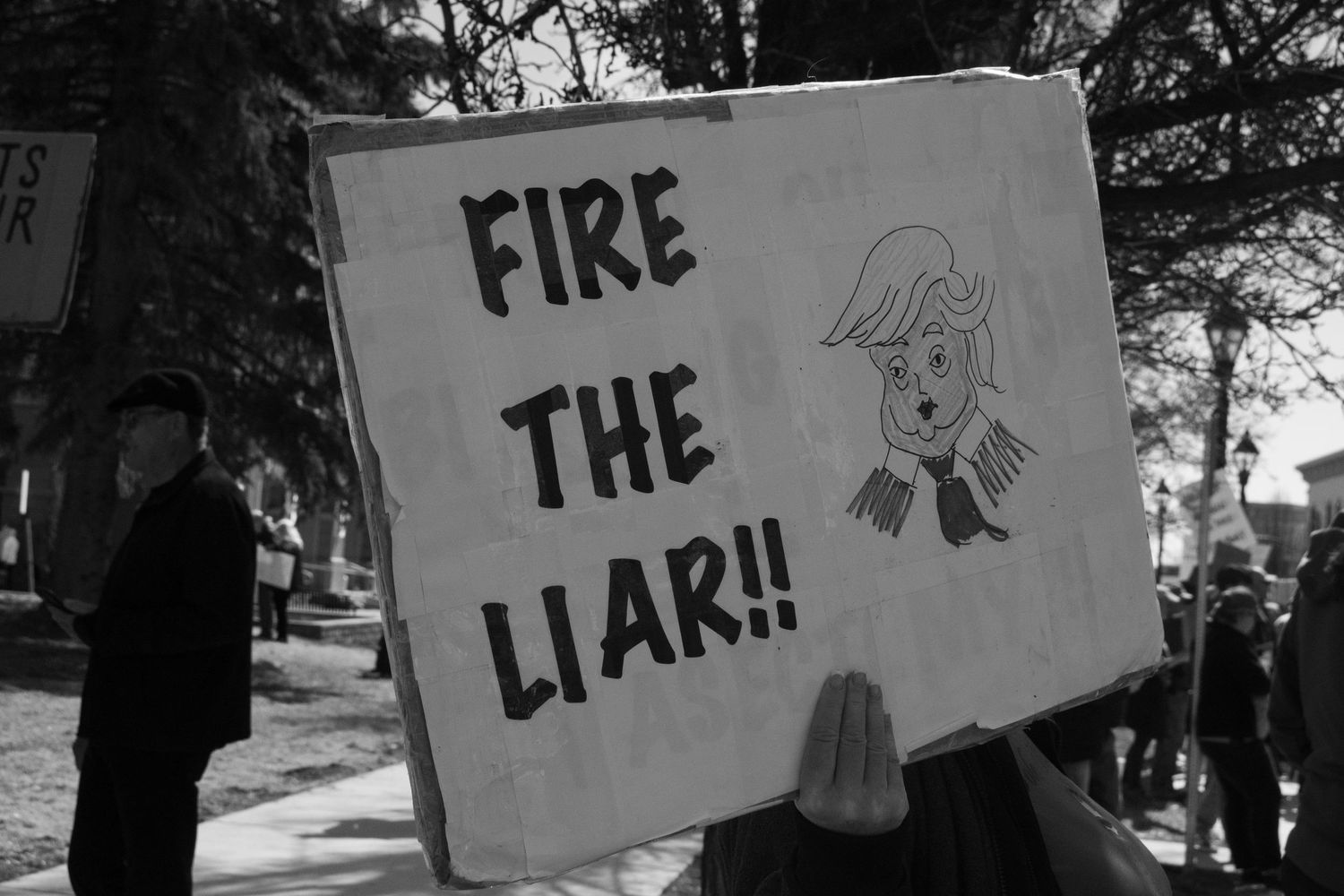 Black and white photo of protest sign reading Fire The Liar with cartoon figure at political rally.