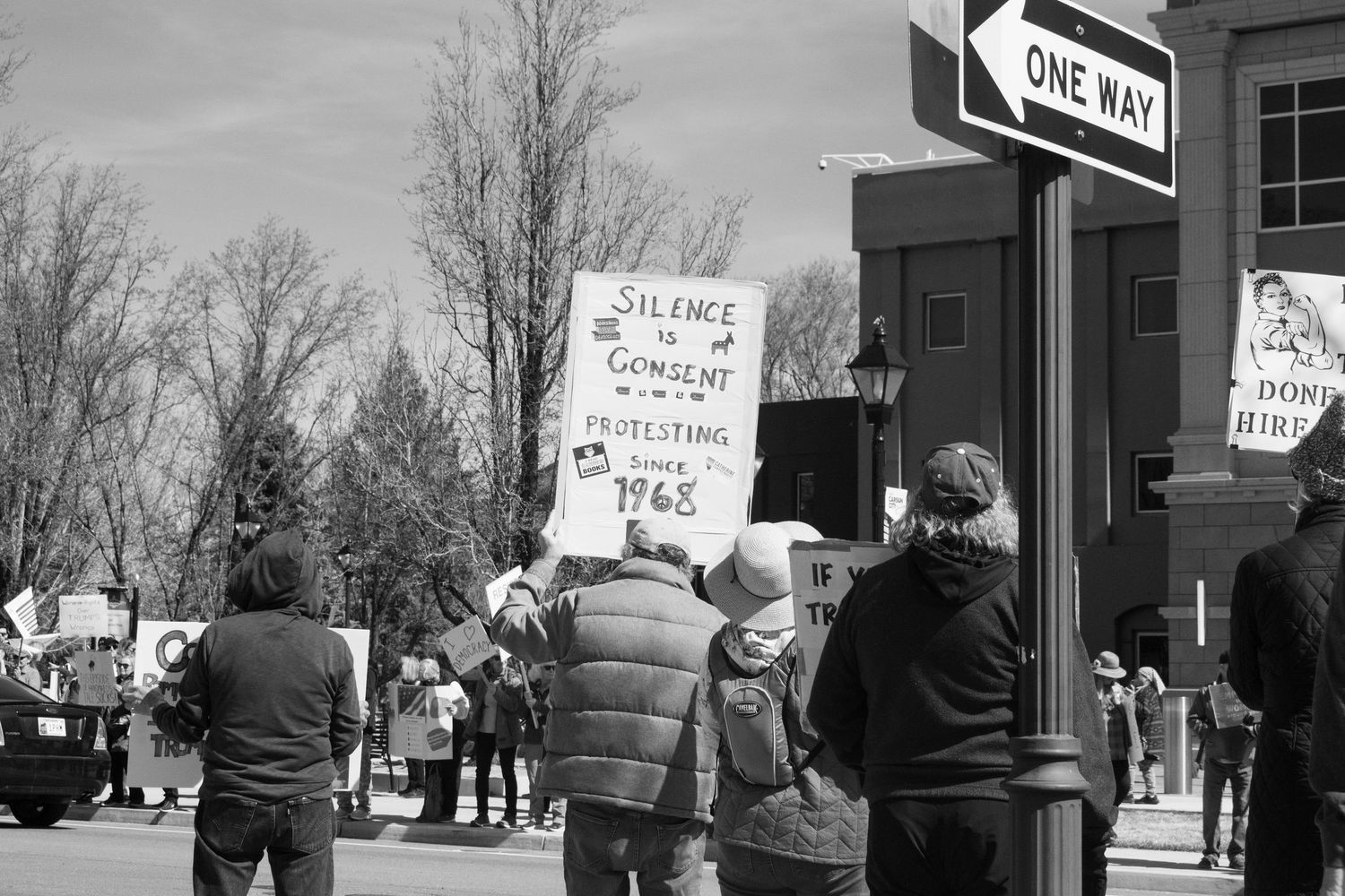 Black and white photo of protesters holding signs including Silence is Consent at a street corner with a One Way sign.