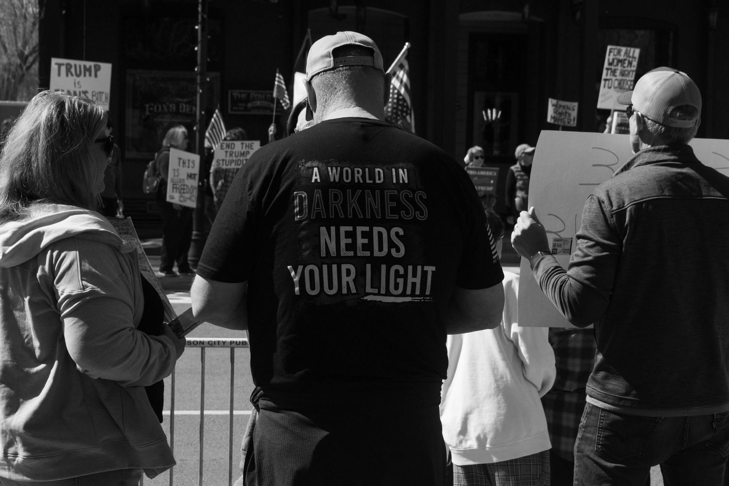 Man wearing A World In Darkness Needs Your Light shirt stands among protesters at outdoor rally.