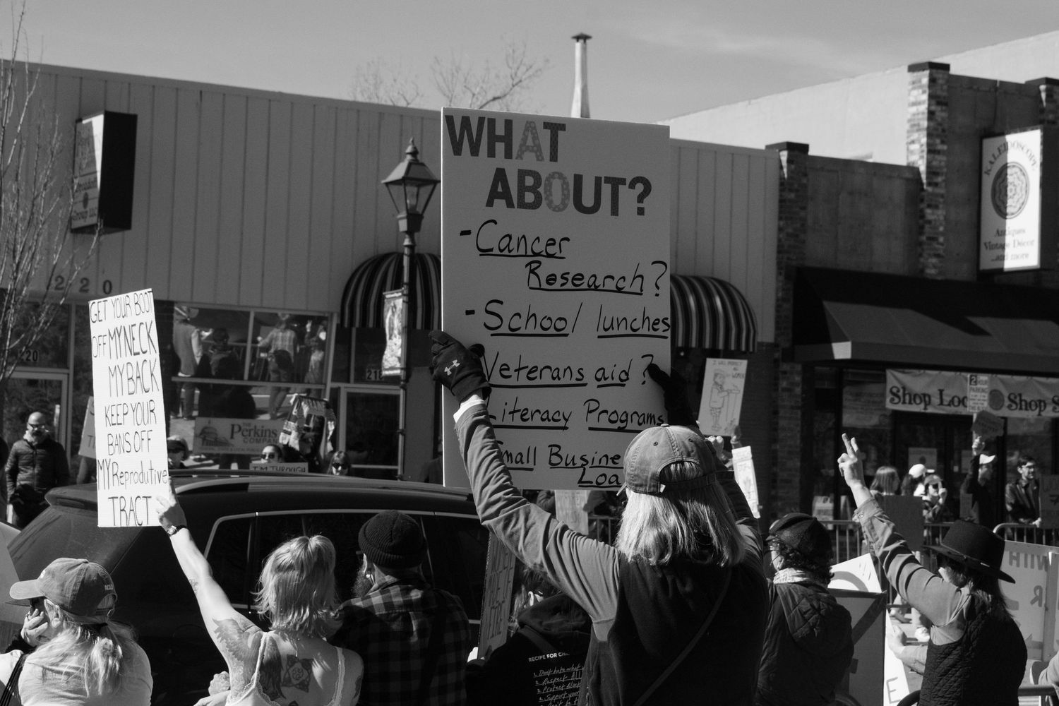 Protester holds sign reading What About Cancer Research School Lunches Veterans at outdoor rally.