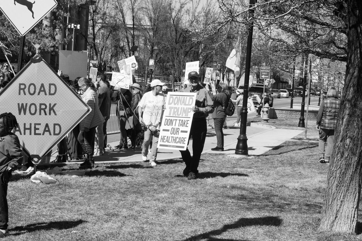 Black and white photo of protesters marching on a sidewalk near a Road Work Ahead sign.