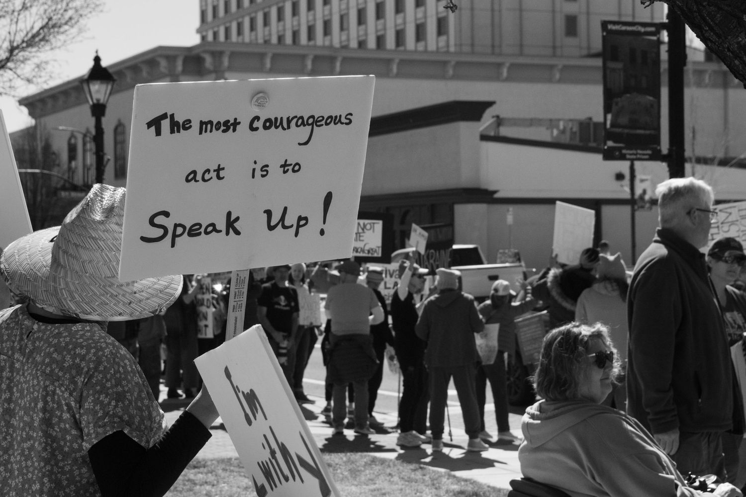 Protester holds sign reading The Most Courageous Act Is To Speak Up at an outdoor rally.