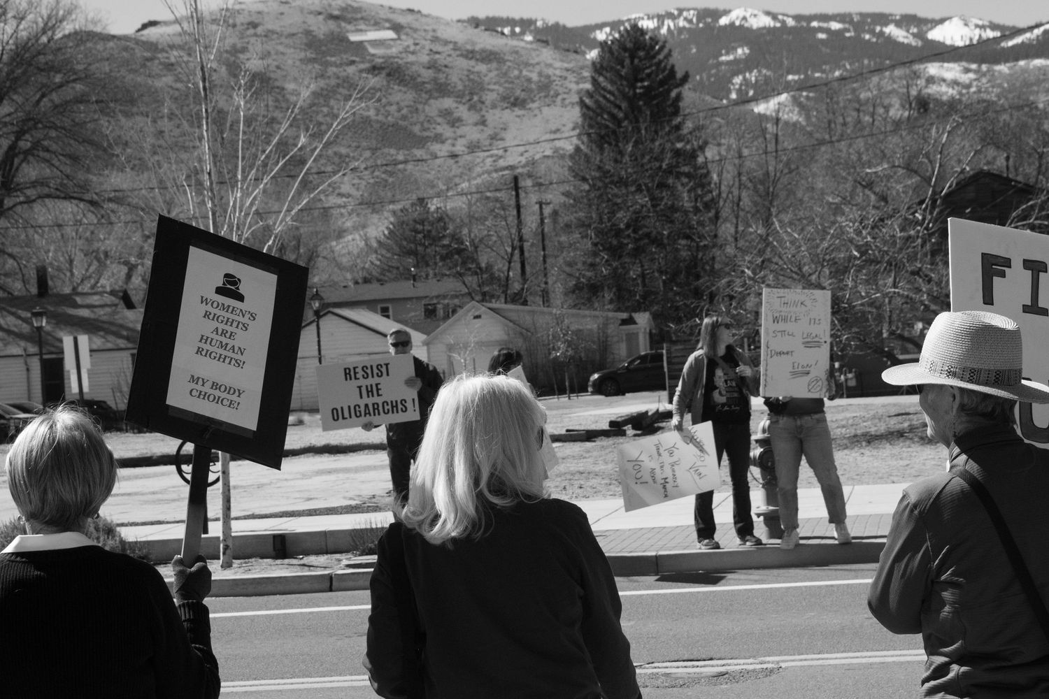 Black and white photo of protesters holding Resist signs along a roadside with snow-capped hills.