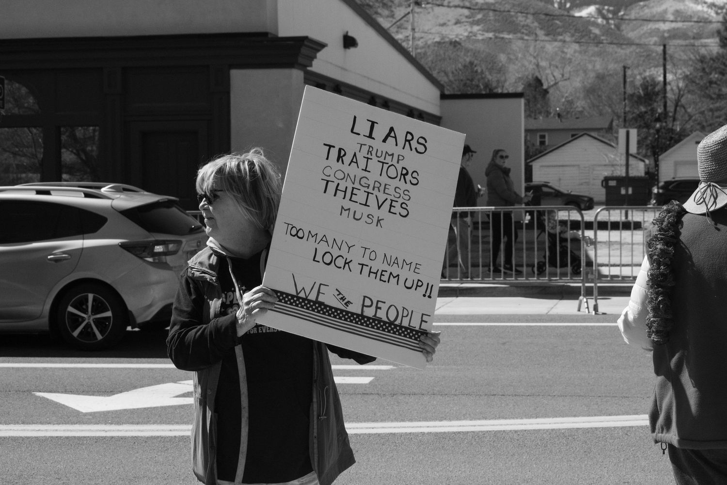 Black and white photo of a woman holding a protest sign reading Liars Traitors Thieves at an outdoor rally.