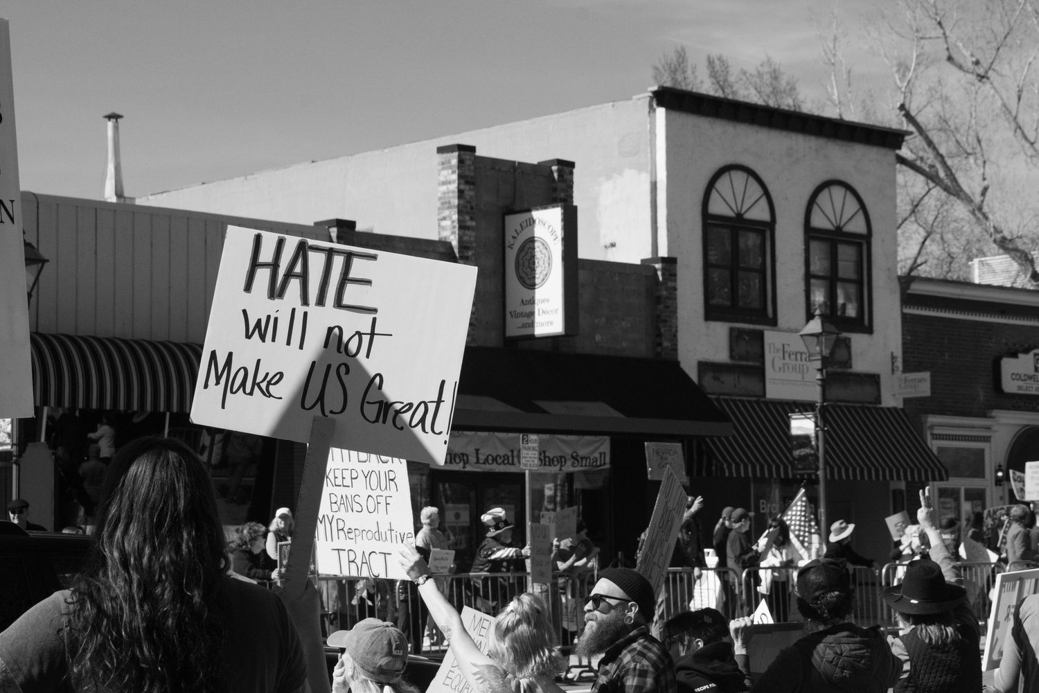 Black and white protest photo with crowd holding a sign reading Hate Will Not Make US Great outdoors.