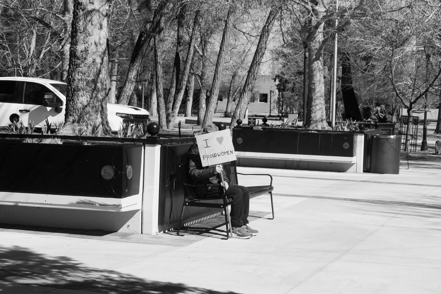 Black and white photo of a lone person sitting on a bench holding a small protest sign in a plaza.