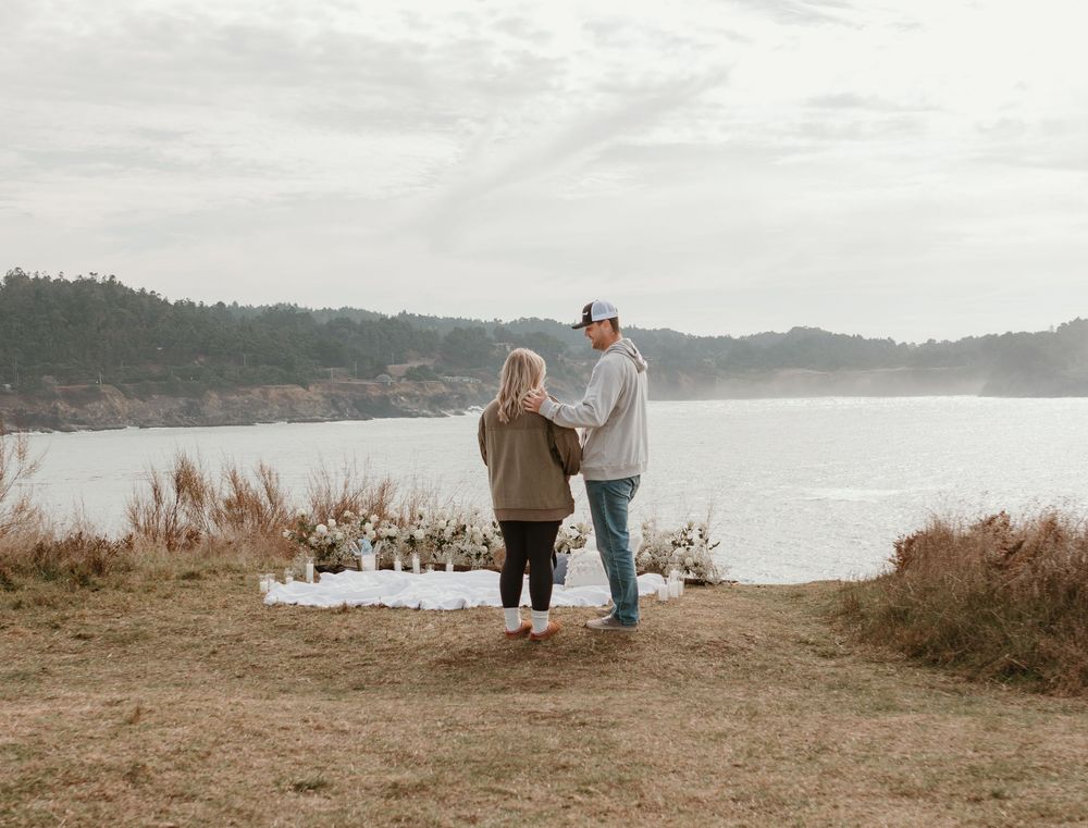 A couple stands on a scenic coastal cliff overlooking a serene bay with mountains in the distance.