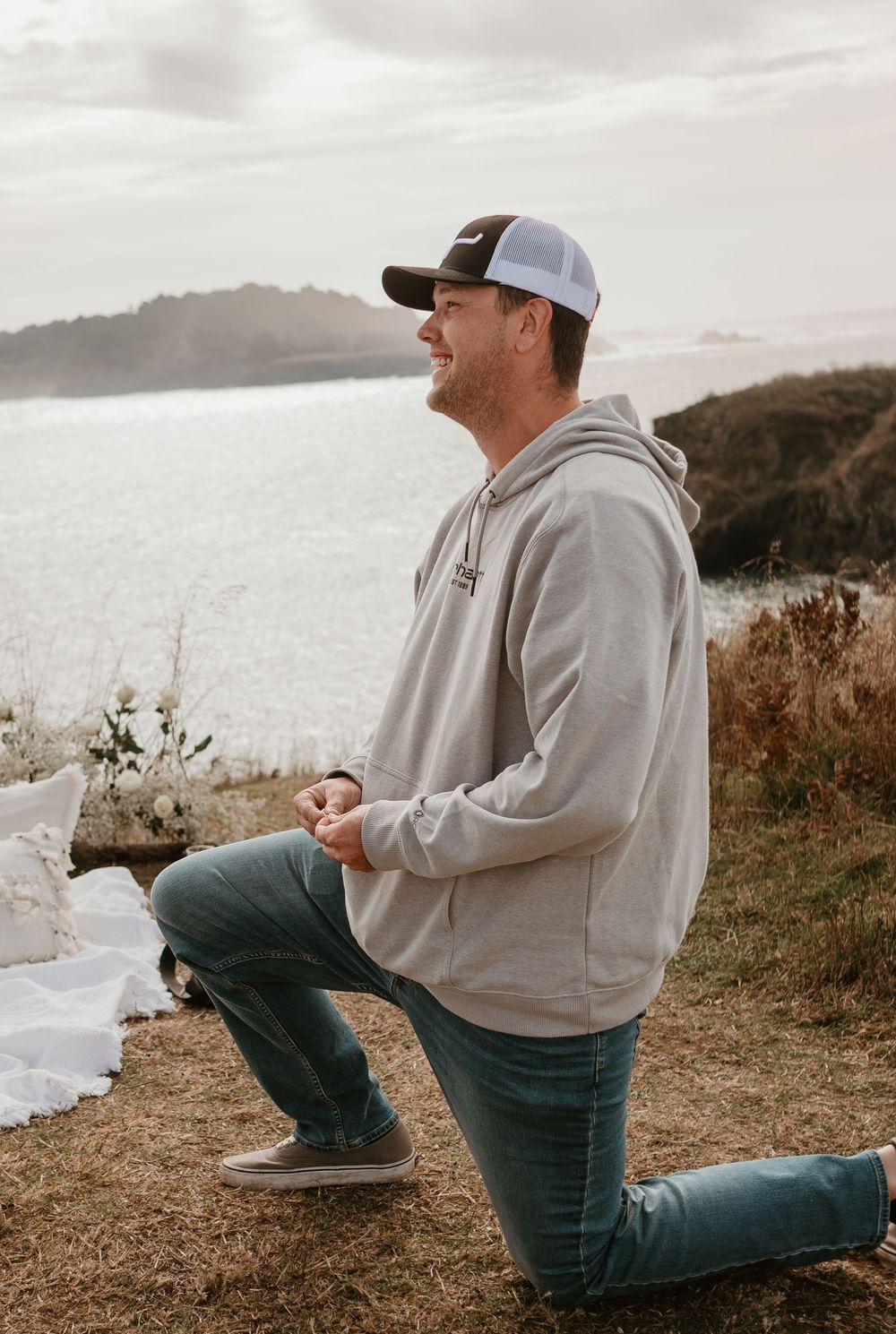 A person kneels down on a scenic coastal overlook with water and mountains in the background.