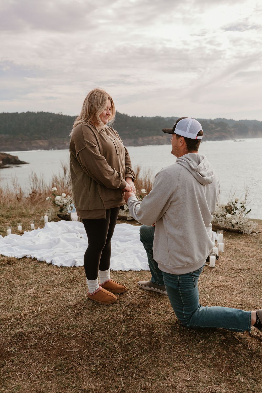 A romantic proposal scene unfolds on a coastal cliff with white flowers and ocean views.