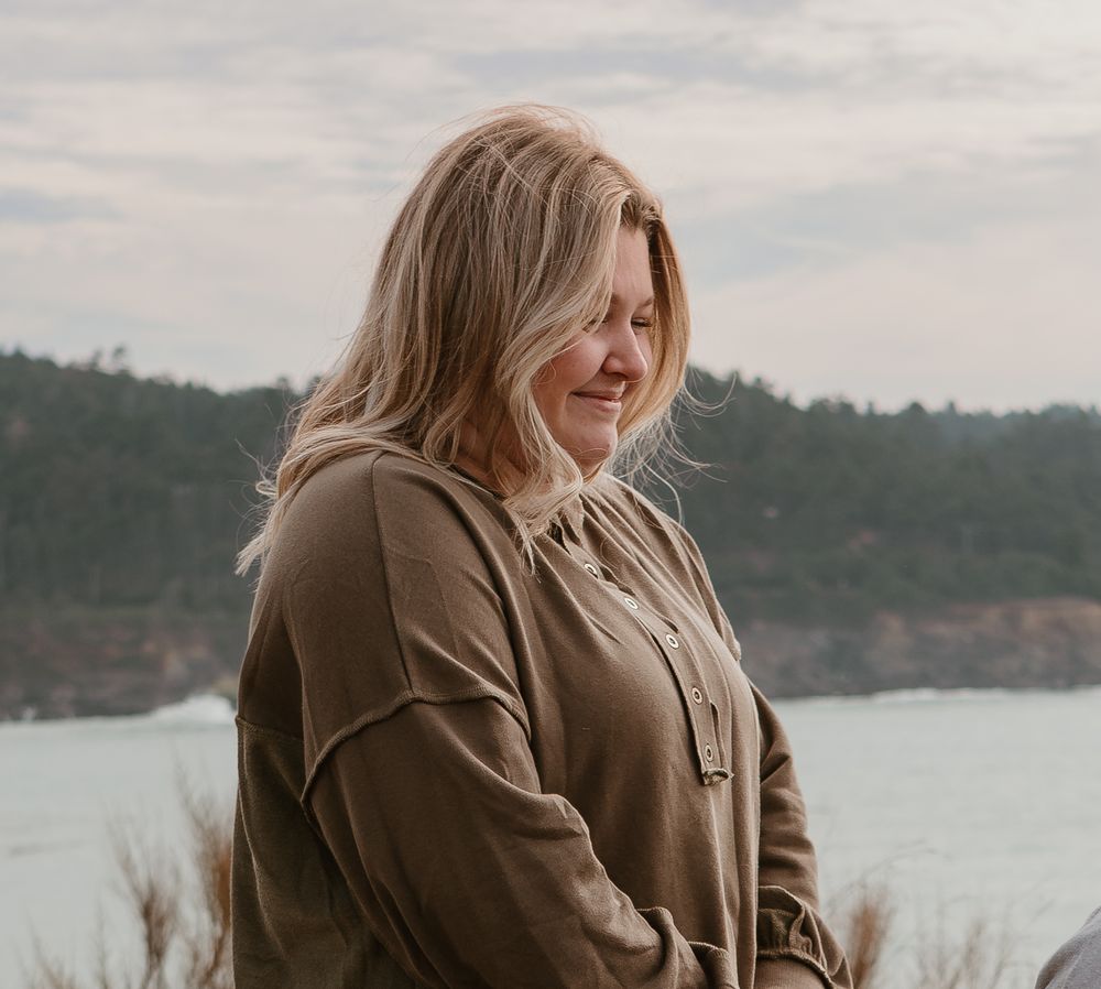 A person in a brown coat stands against a cloudy coastal landscape backdrop.