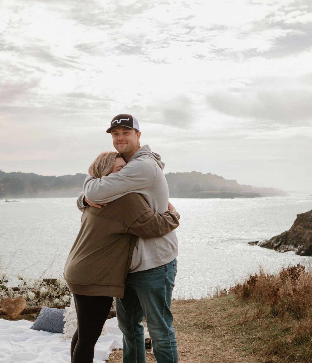 Two people share a heartfelt embrace on a coastal cliff with mountains in the background.