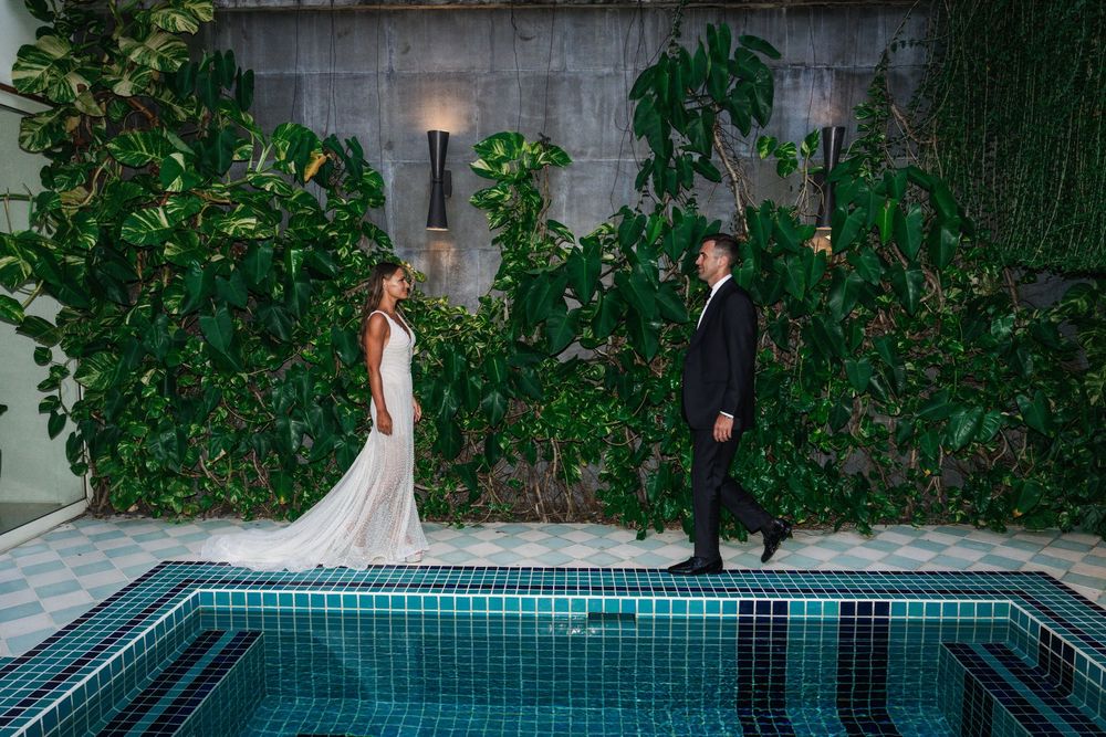 A couple shares a romantic moment beside a turquoise pool surrounded by lush greenery at dusk.