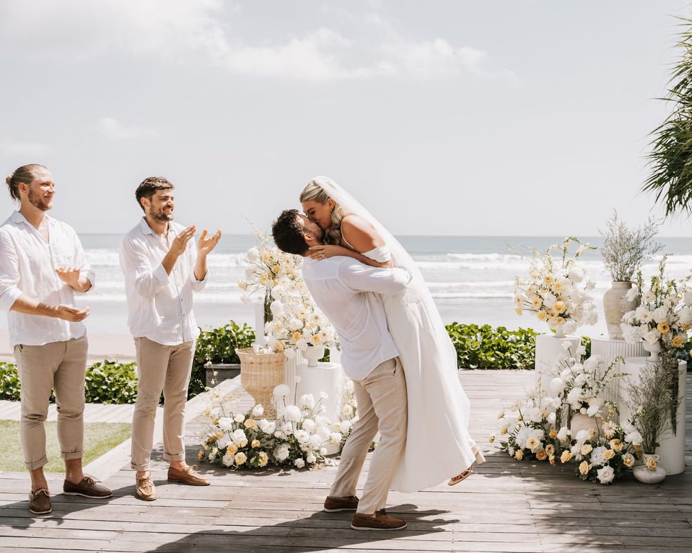 Wedding couple in all white beachfront celebration with tropical floral arrangements at beachside wedding ceremony.