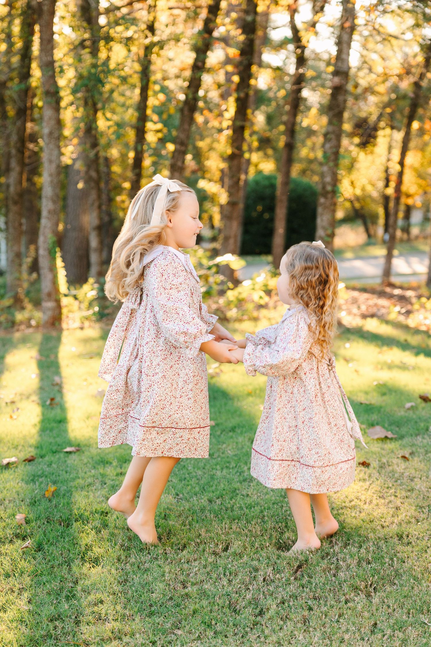 Two figures in matching floral dresses dance and twirl together in a sunlit wooded area during golden hour.