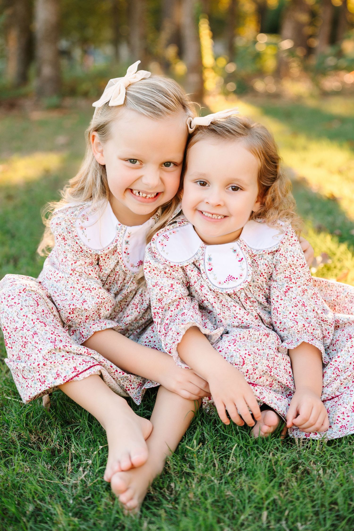 In the autumn sunlight, three siblings wearing matching floral dresses lie together playfully on green grass.