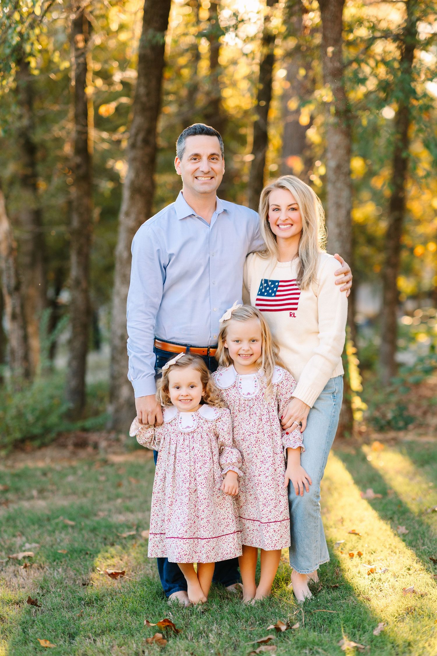 A family wearing coordinated blue and floral outfits poses in a sunlit forest setting during golden hour.