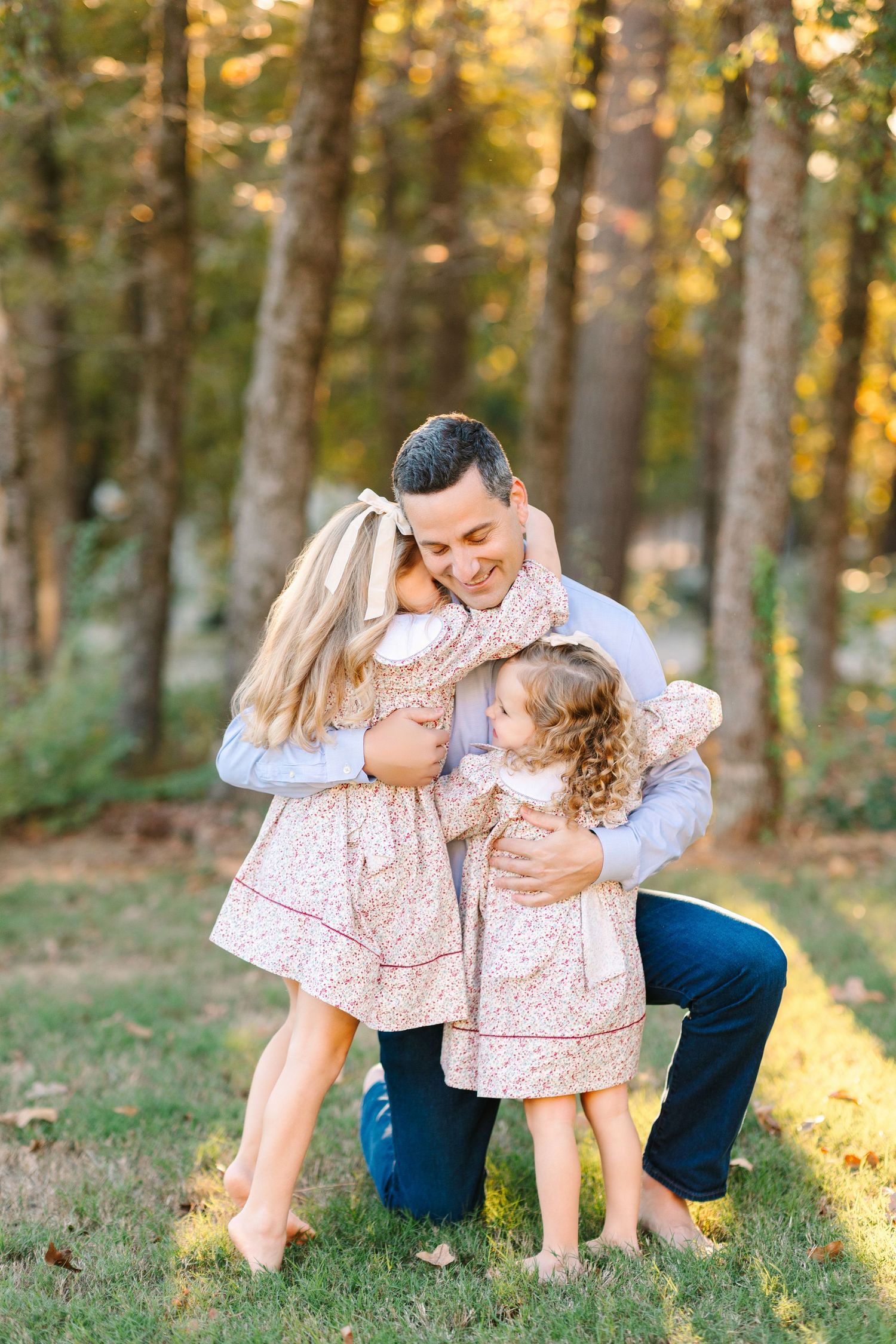 A tender family moment shared in a sunlit forest with two young sisters in matching pink dresses hugging their parent.