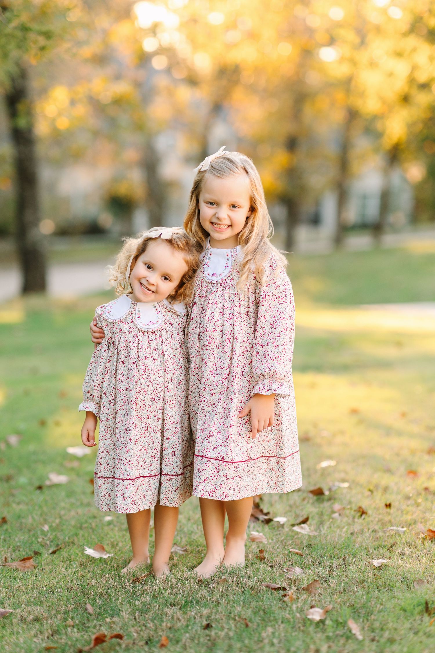 Two children wearing matching floral dresses stand together on grass during autumn golden hour.