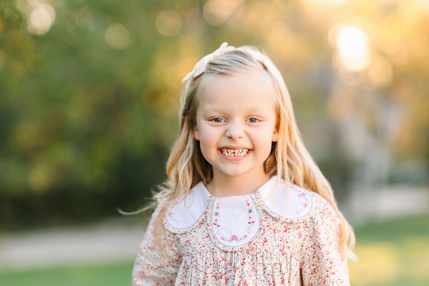 A cheerful young child with blonde hair wearing a floral dress with a peter pan collar in a sunny outdoor setting.