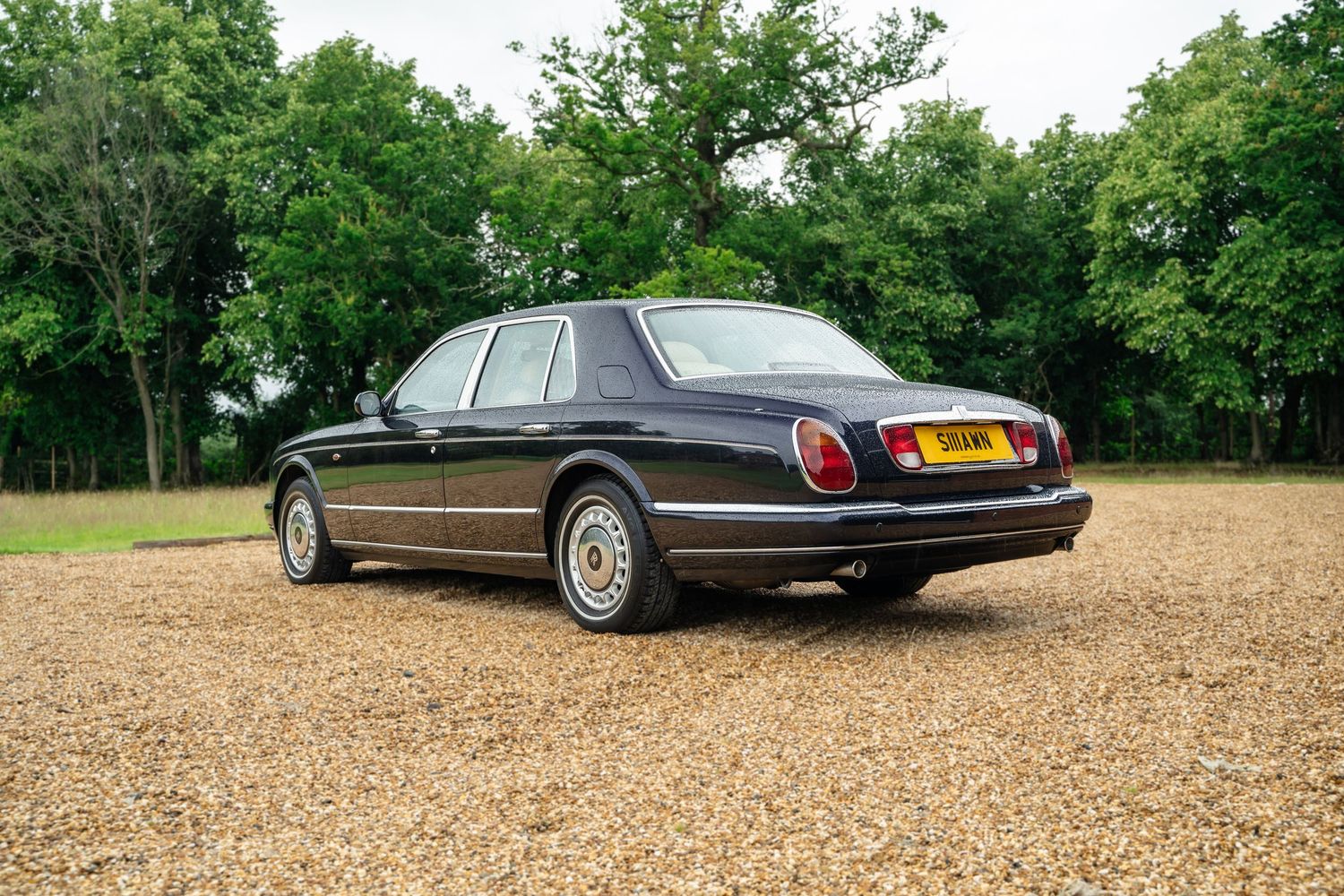 Elegant black luxury cars displayed on gravel lot near wooded area.
