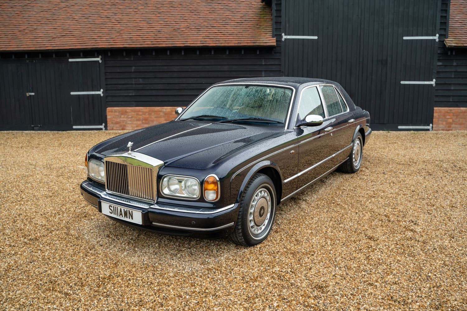 Classic luxury cars parked outside a black wooden barn with gravel parking area.