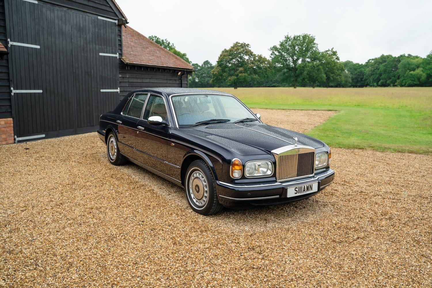 Side view of vintage luxury vehicles displayed on gravel near black barn buildings.