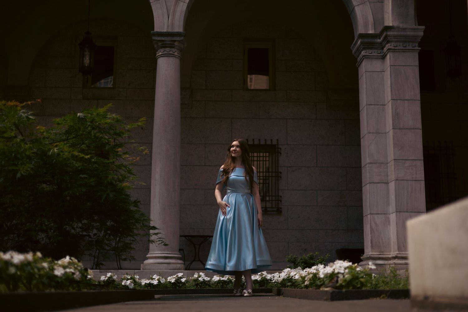 A person in a flowing blue dress stands in an elegant stone archway surrounded by white flowers.