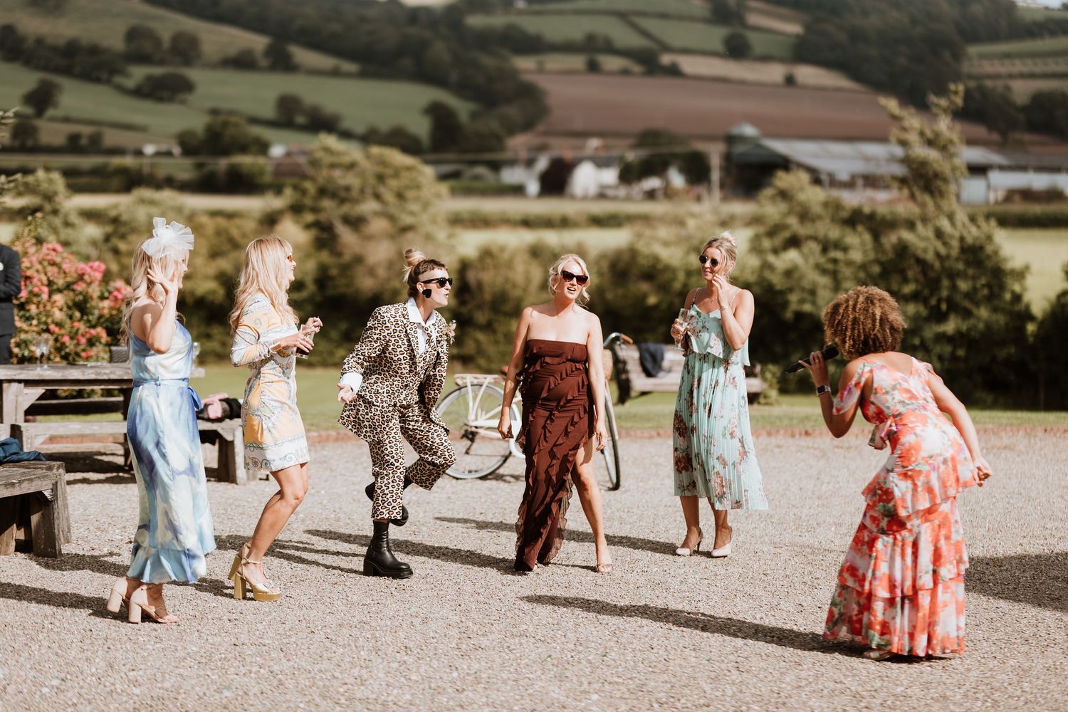 Group of guests in summer clothing dance outdoors on a sunny day with rolling hills in the background during drinks reception