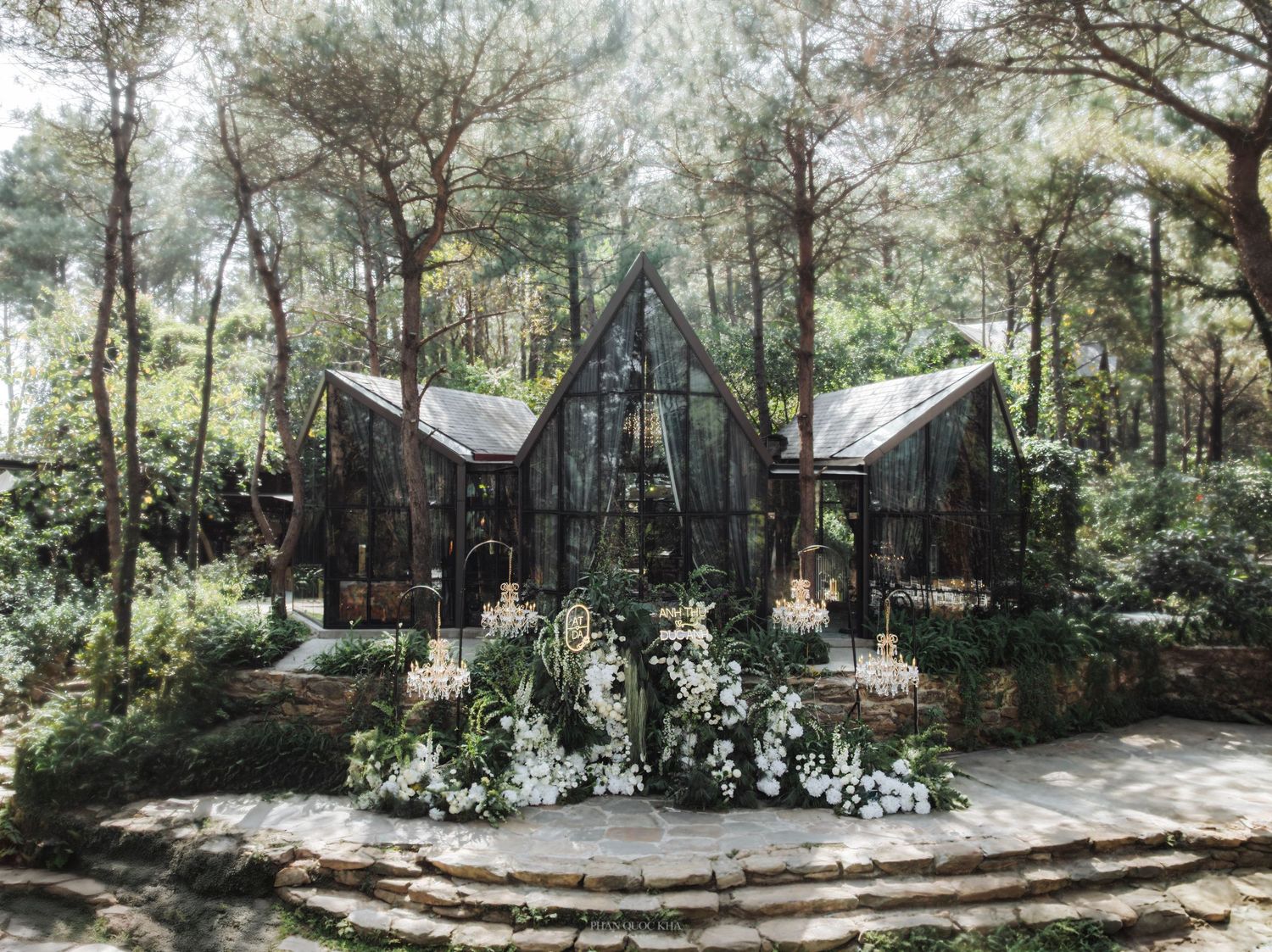 Stunning glass chapel wedding venue surrounded by tall pine trees with white floral arch decorations.