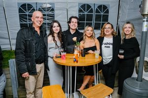 Group of friends enjoying drinks at outdoor bistro tables near a grey wall in casual evening attire.