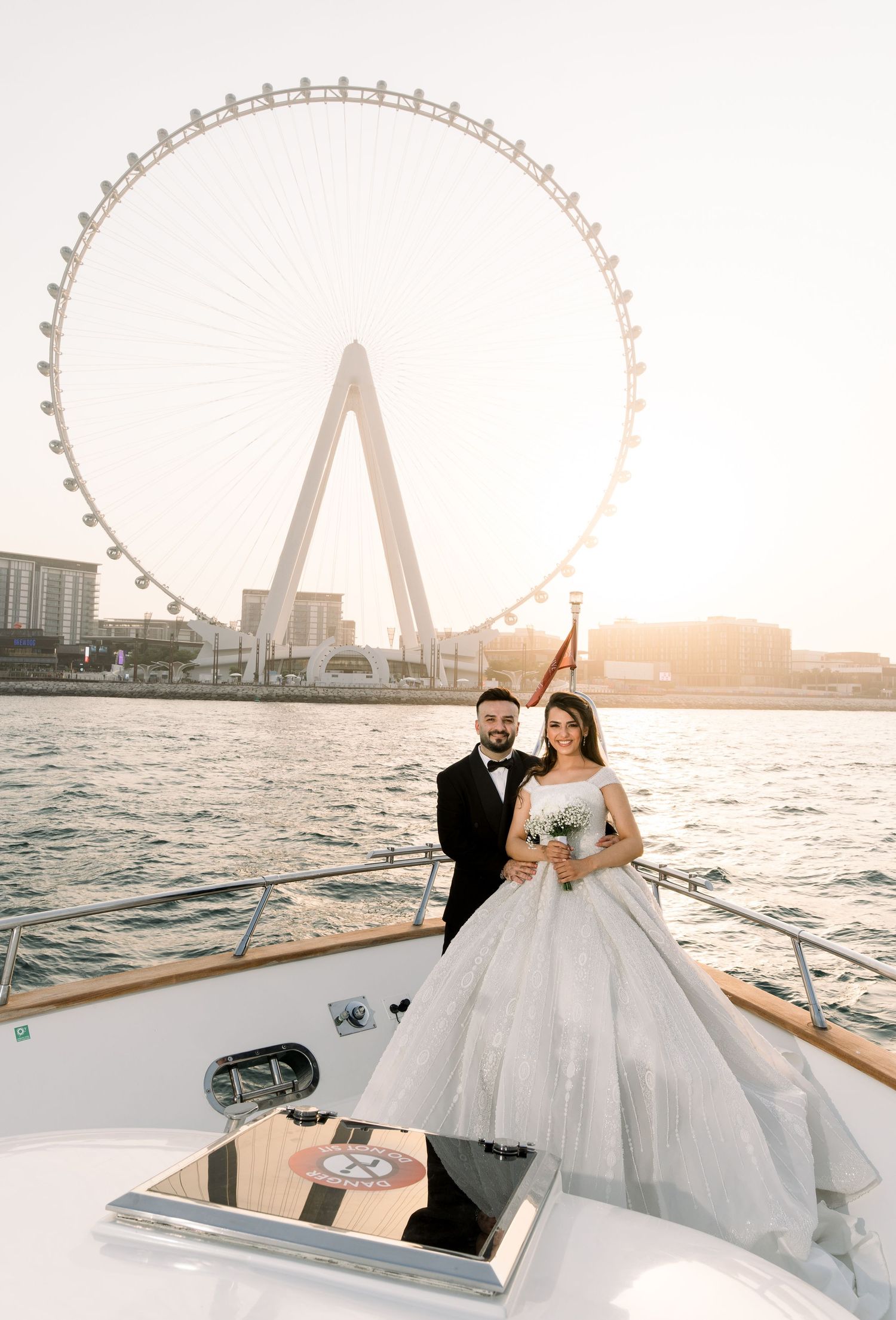 A romantic couple posing on a luxury yacht with Dubai Eye observation wheel visible in the background at sunset.