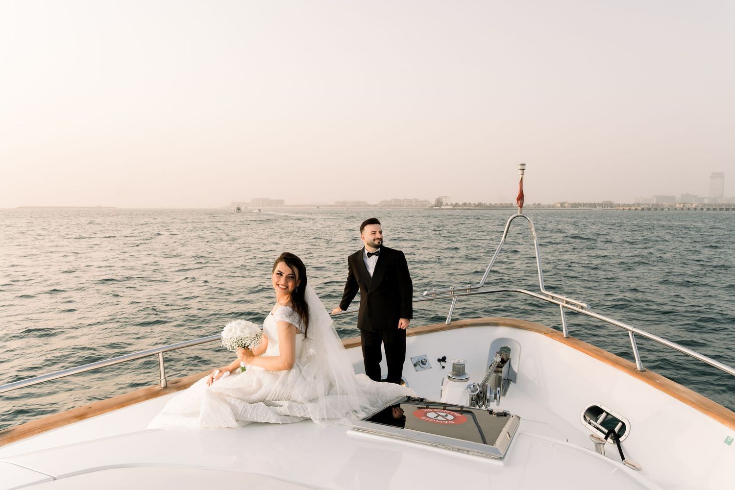 Wedding couple sits on bow of luxury yacht during romantic sunset cruise on calm ocean waters.