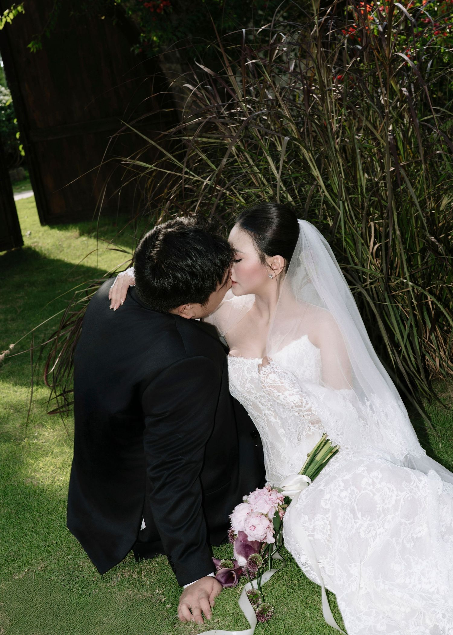 Bride and groom share romantic kiss in garden, bride holding pink floral bouquet.