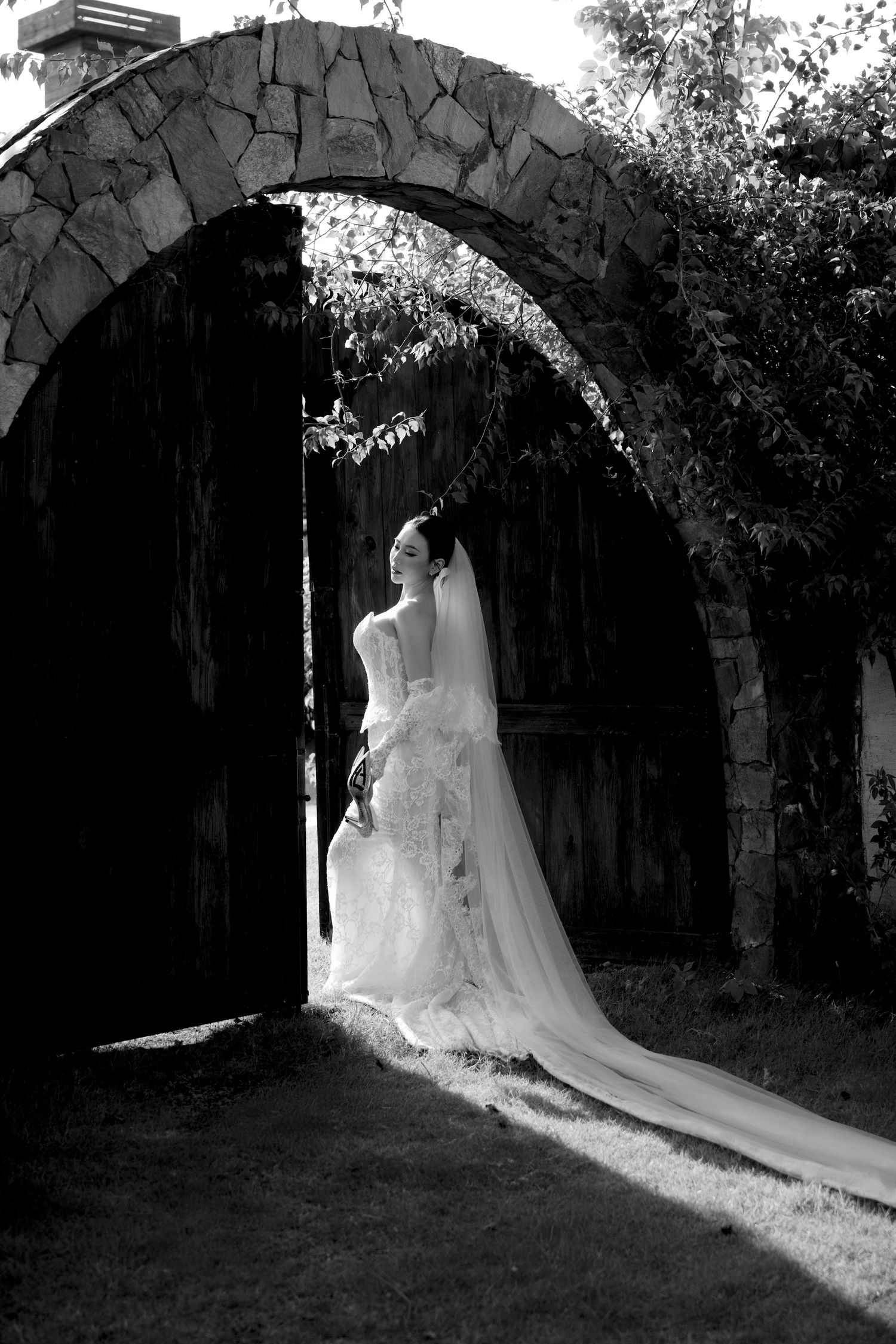 Black and white photo of a bride in a flowing lace gown and veil standing beneath a rustic stone archway.