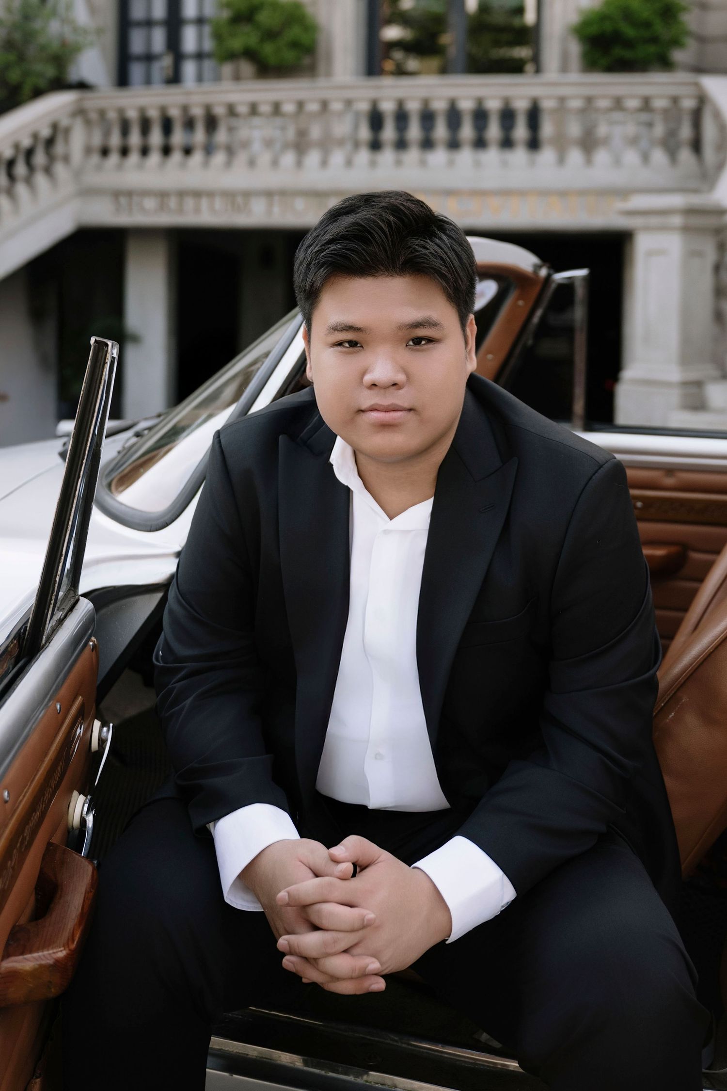 Young Asian groom in black tuxedo sits confidently beside vintage car at Secretum Hotel.