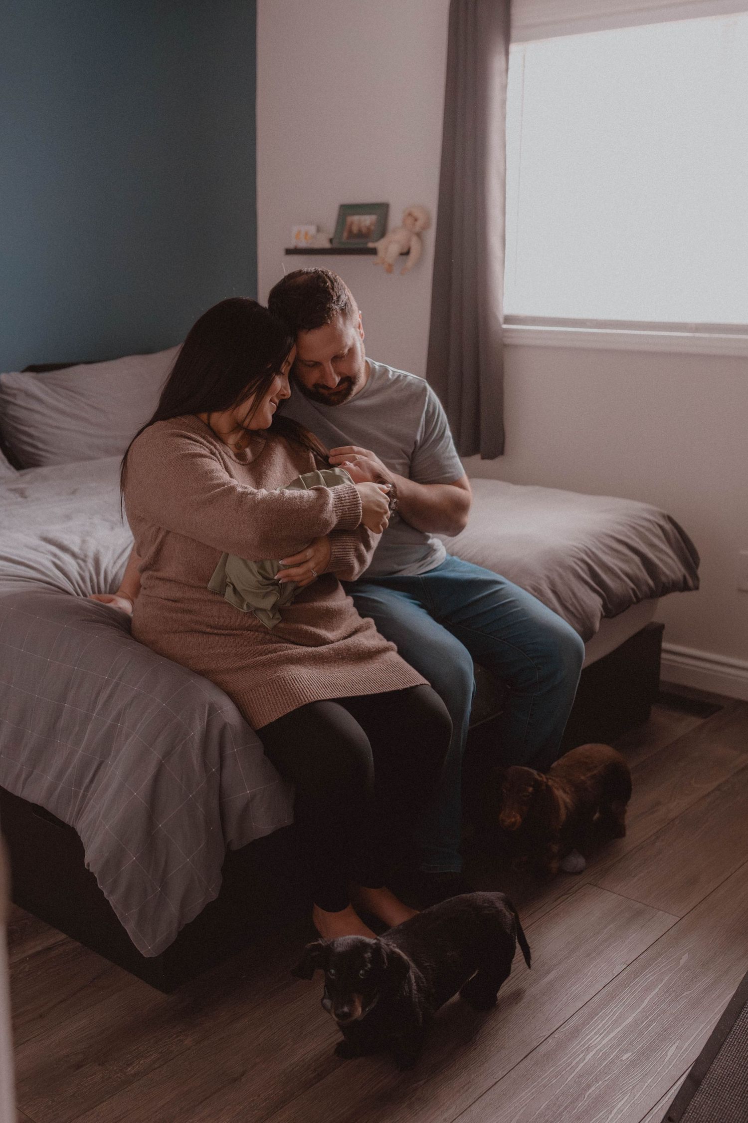 Couple sitting on bed holding newborn baby with two dachshund dogs on floor in cozy bedroom.