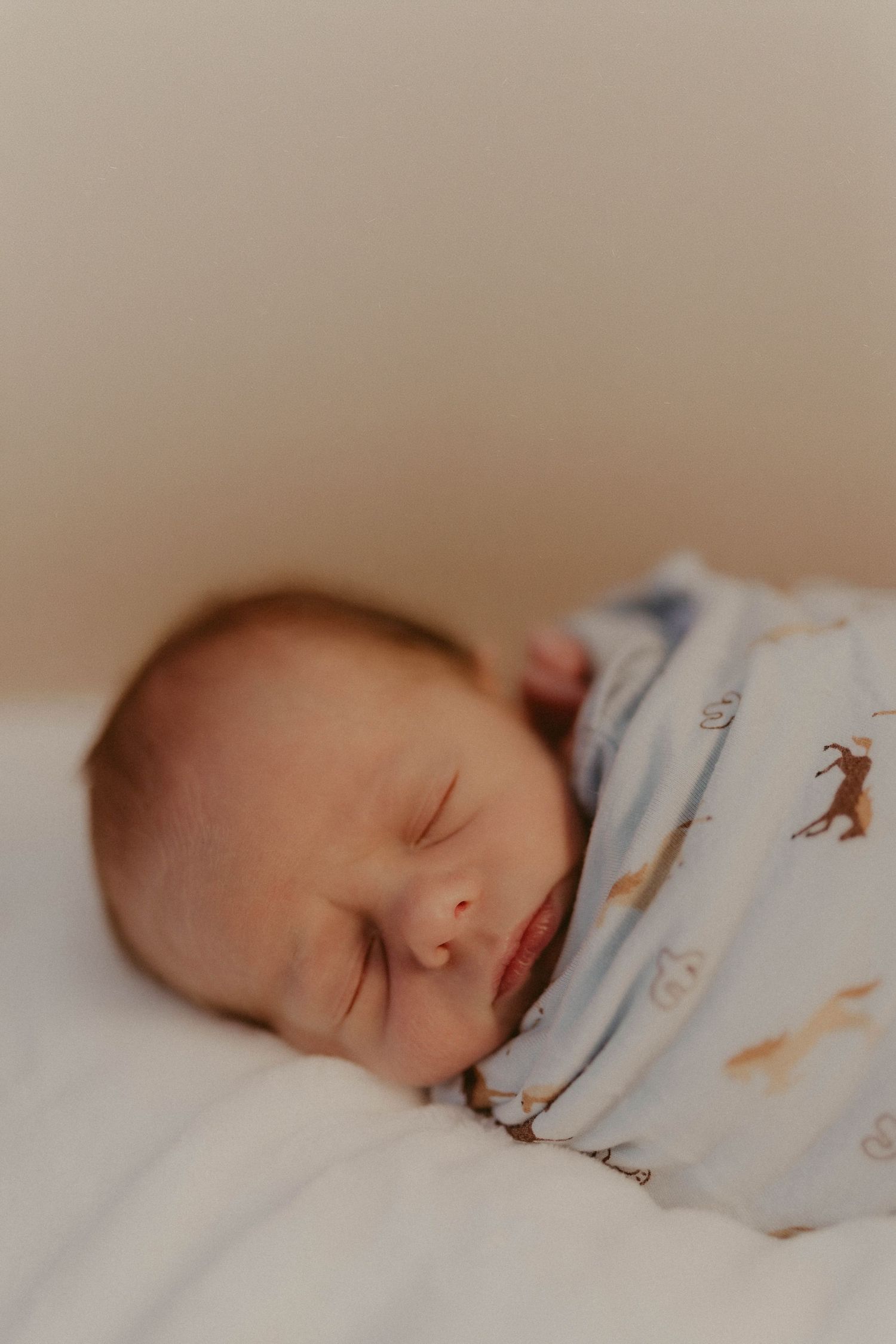 Peaceful newborn baby sleeping on white bedding, wearing a white swaddle with animal prints.
