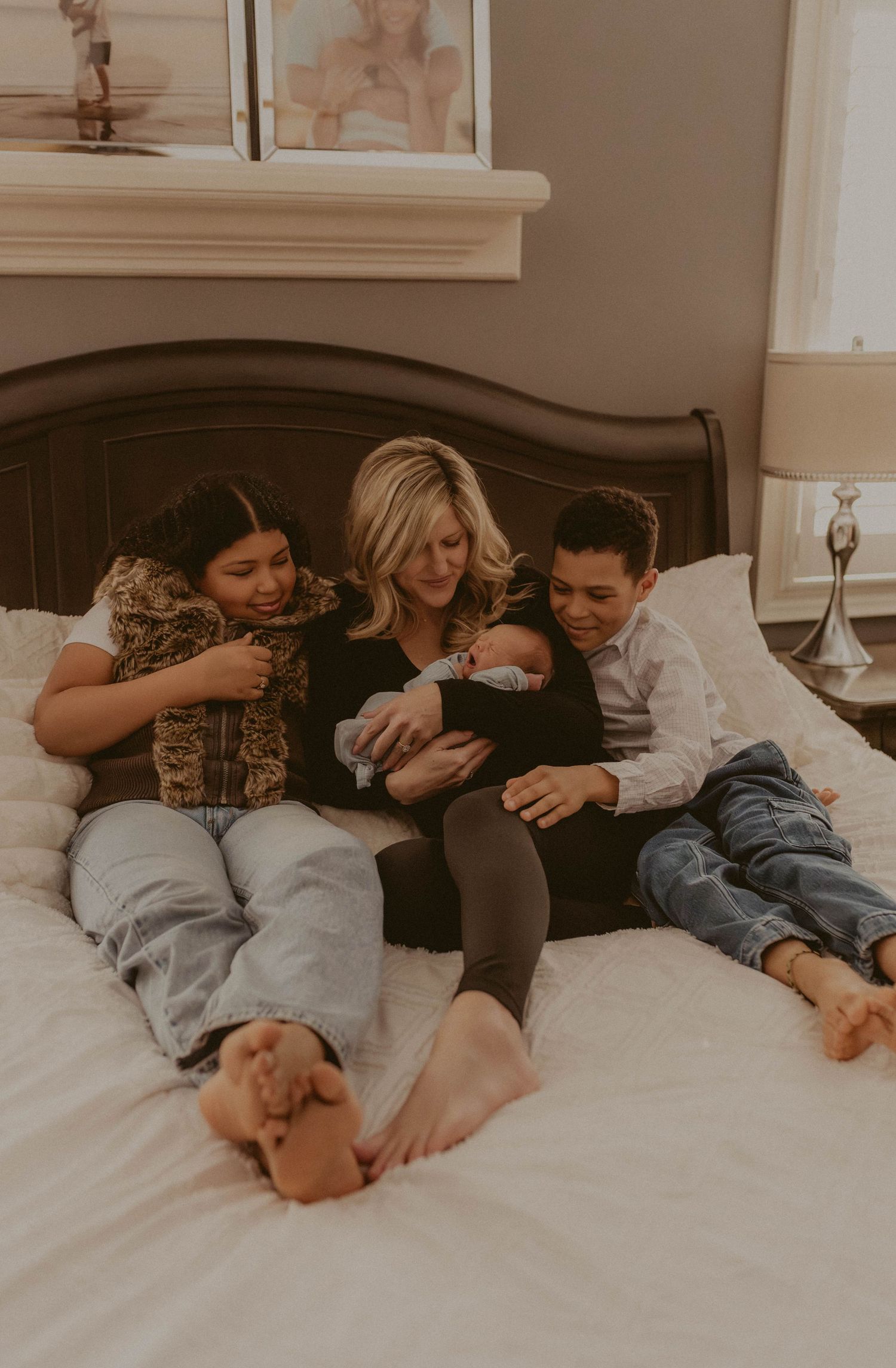 A blonde mother sits on a white bed holding a newborn, surrounded by her two older children in a warm family portrait.