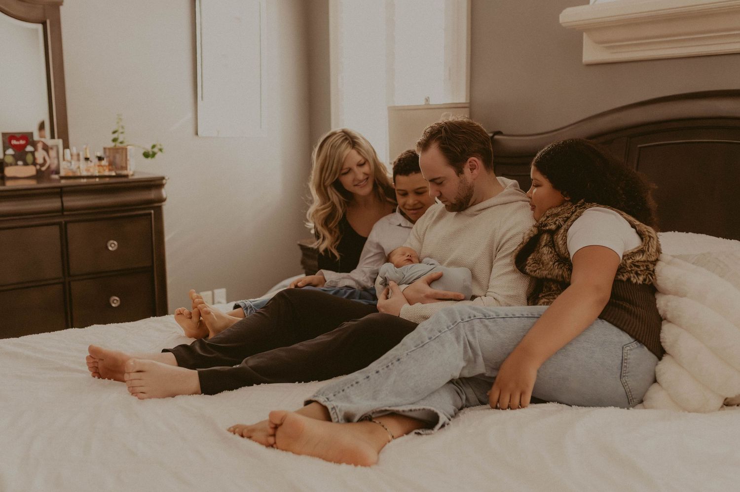 Family of four relaxing on a white bed, smiling and bonding with their newborn baby in a cozy bedroom.