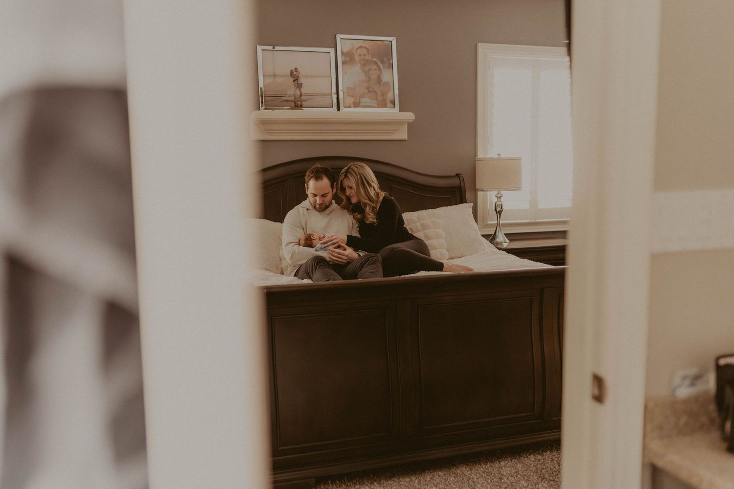 Couple sitting together on a dark wood bed, looking at their newborn baby in a warmly lit bedroom.