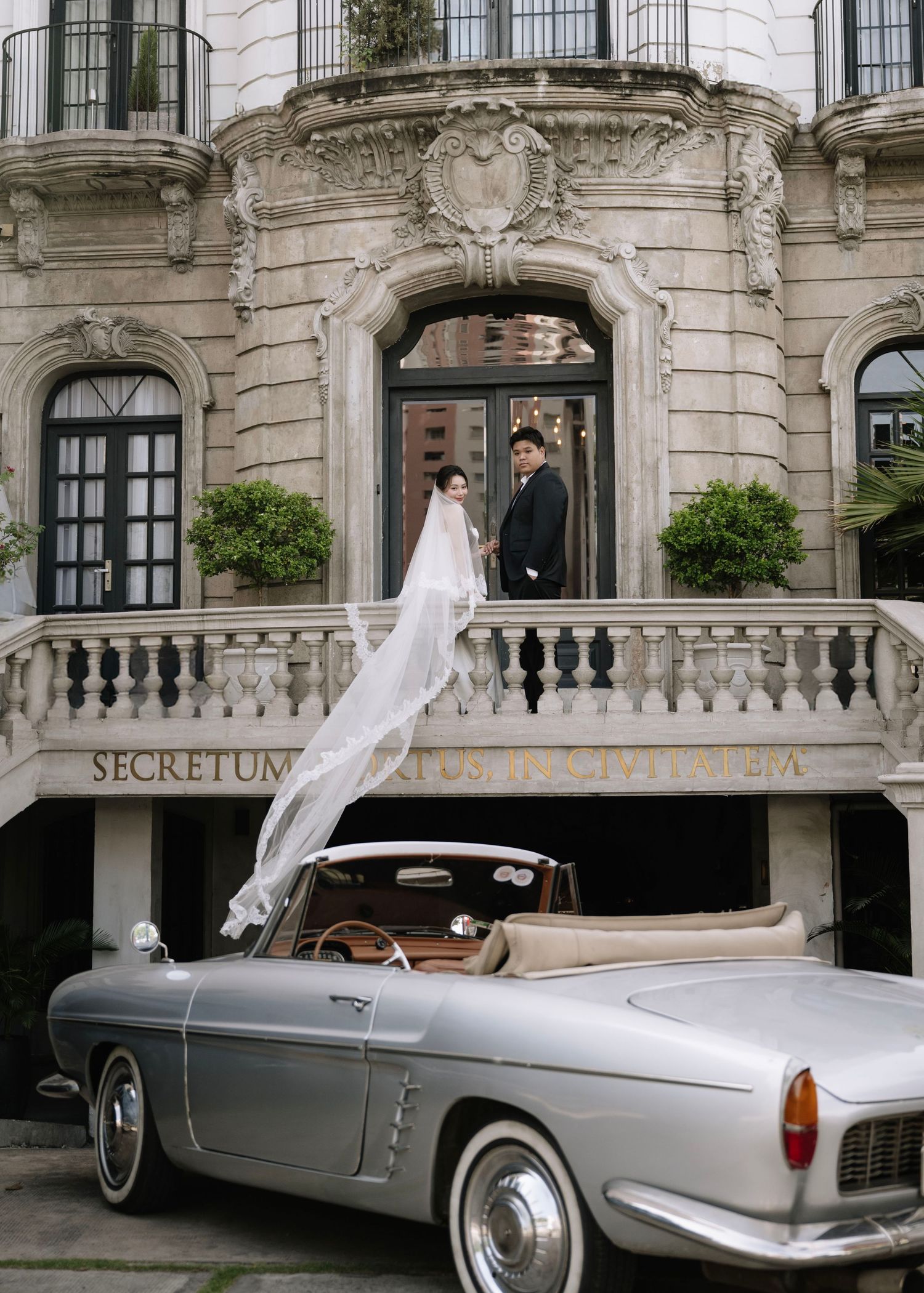 Wedding couple stand on balcony of ornate building above a classic silver vintage convertible car.