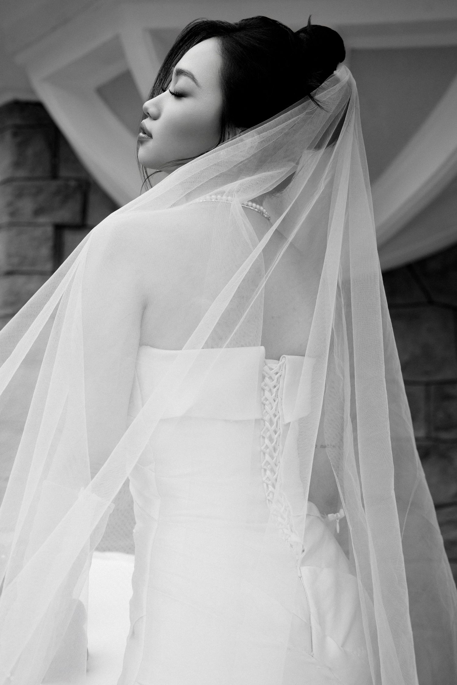 Black and white portrait of Asian bride in strapless wedding dress with long flowing veil from behind.