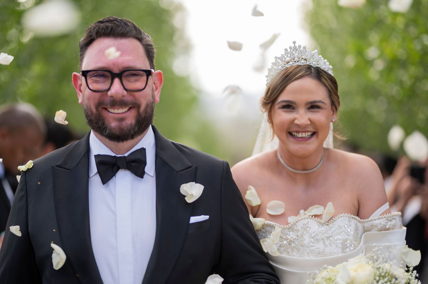A bride and groom share a joyful moment during their outdoor wedding ceremony surrounded by falling flower petals.