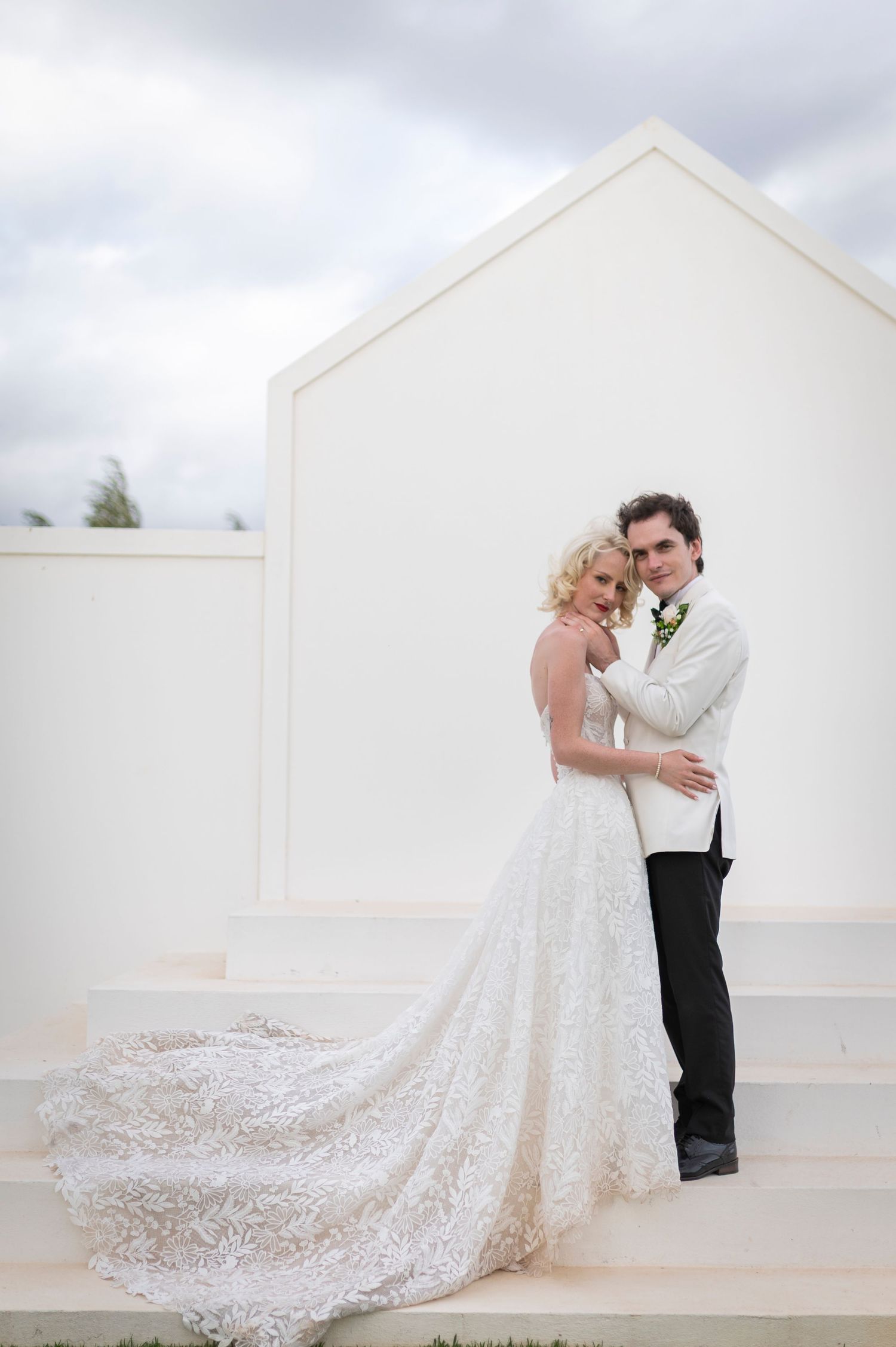 A romantic wedding couple embraces on a white rooftop terrace with a flowing lace bridal gown train.