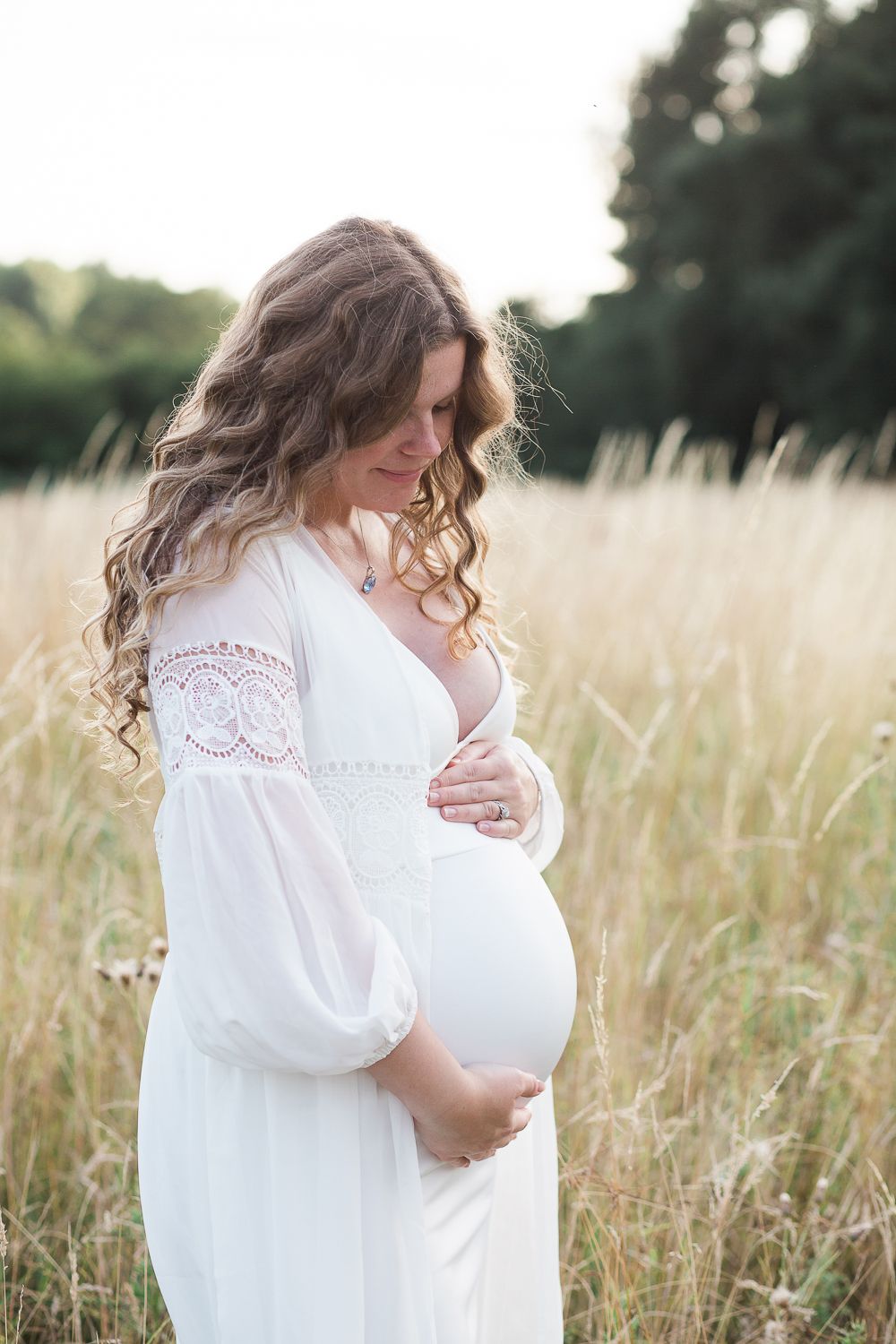Woman in white dress cradles her pregnant belly in sunlit meadow.