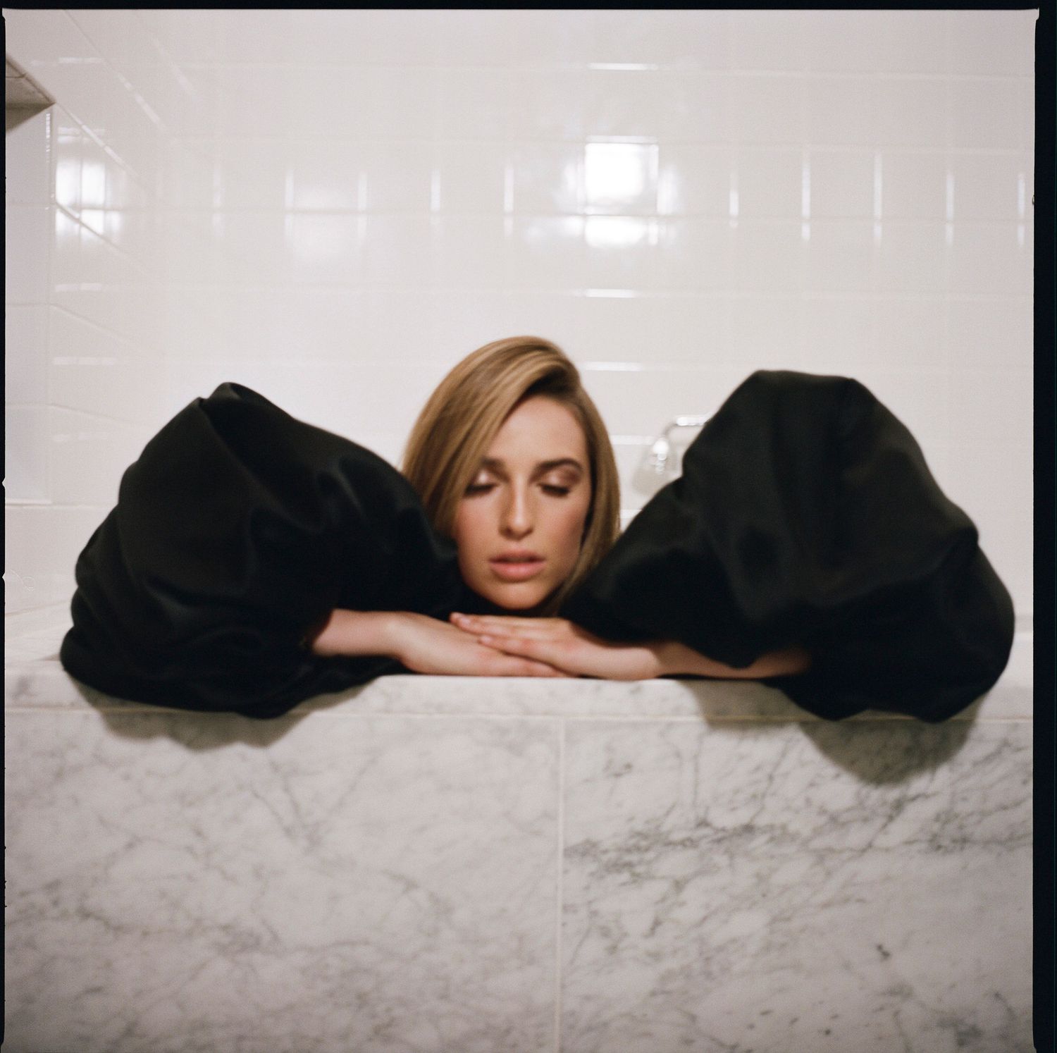 A woman in a black dress rests her head on a marble bathtub edge in a white tiled bathroom.