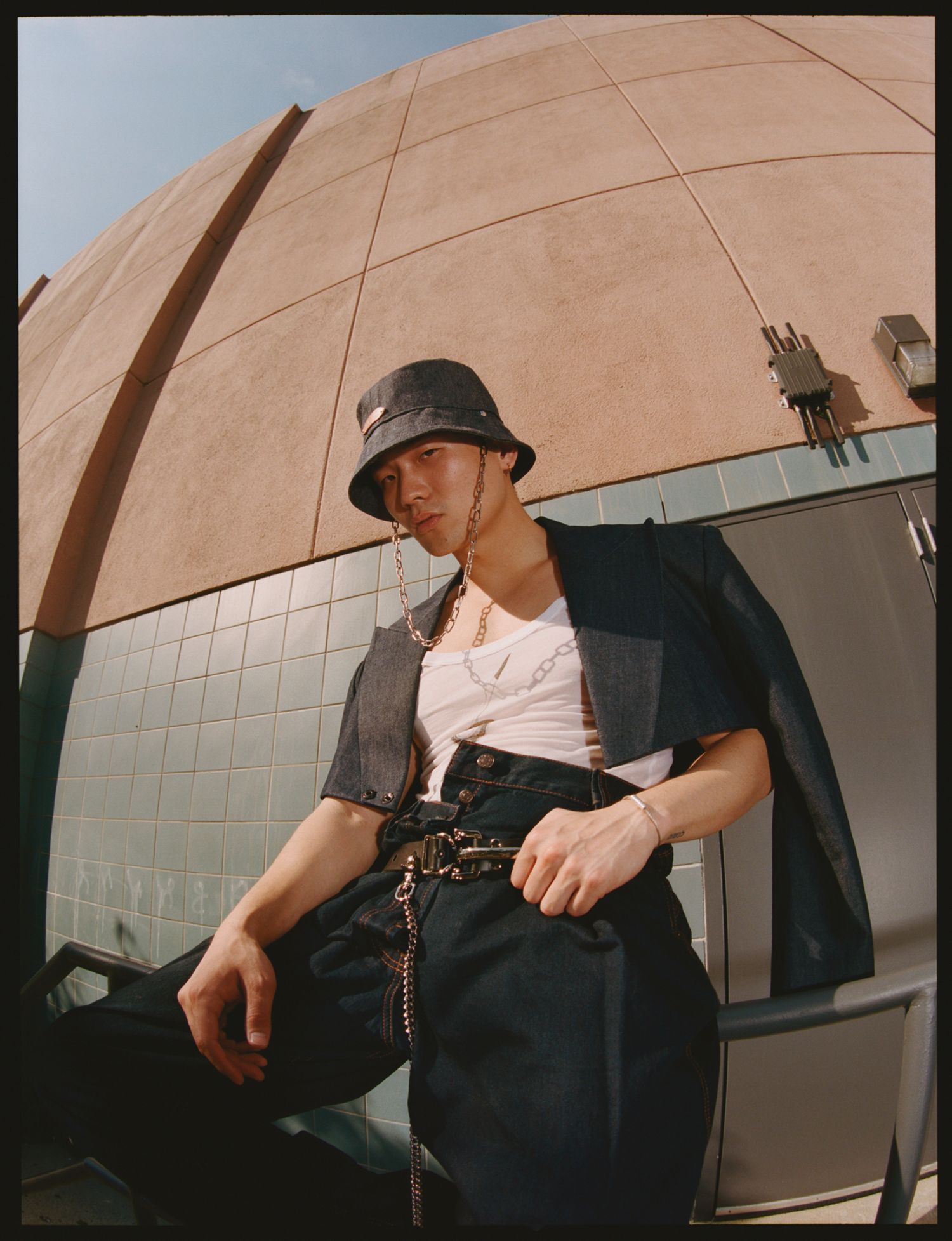 Young man in bucket hat, white tee, dark vest and wide-leg pants poses outdoors near a domed building.